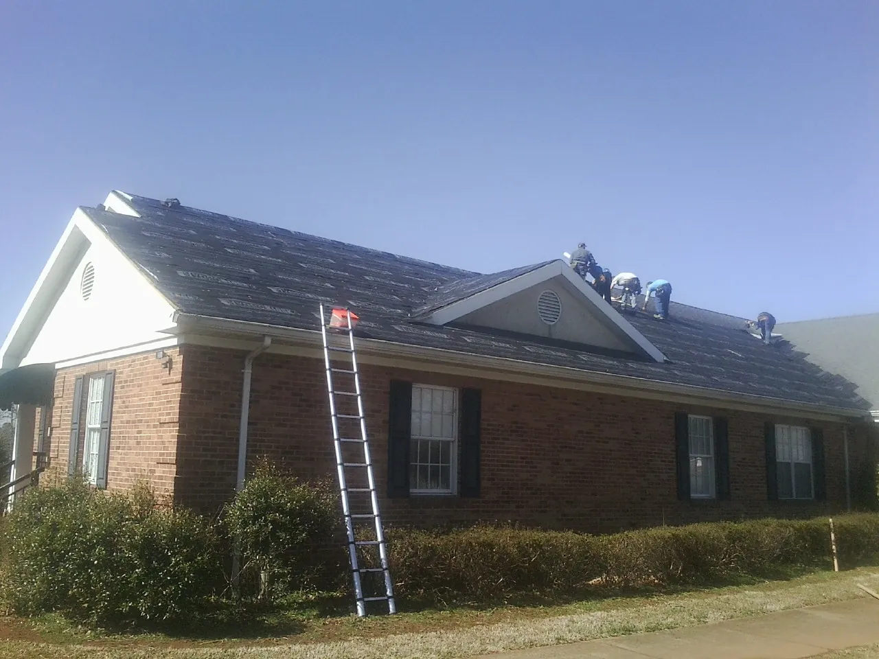 Crew tearing shingles off a brick ranch roof with a ladder set — Metal Roof Repair work by our crew in Flushing
